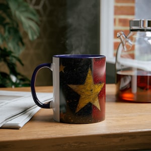 May include: A white ceramic mug with a blue rim and a vintage-style design featuring a large yellow star on a red, white and blue background. The mug is filled with a steaming beverage.