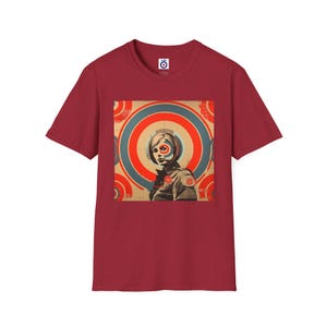 Pode incluir: Uma camiseta vermelha com uma estampa gráfica de uma mulher com um design de alvo atrás dela. O design do alvo é em vermelho, branco e azul.