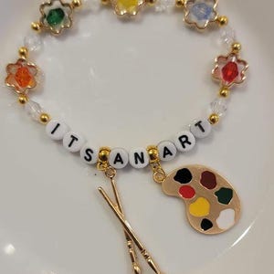 May include: A gold beaded bracelet with colorful flower charms and the words "IT'S AN ART" spelled out in white beads. The bracelet also has a gold paint palette charm and two gold paintbrush charms.