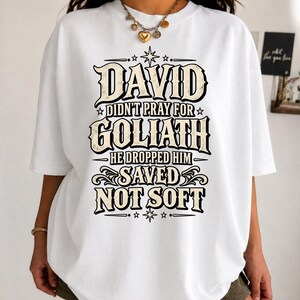 Peut inclure: Un t-shirt blanc à manches courtes avec un texte graphique noir et beige indiquant : DAVID DIDN'T PRAY FOR GOLIATH HE DROPPED HIM SAVED NOT SOFT, agrémenté d'étoiles et de motifs décoratifs centrés sur le devant du vêtement.