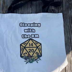 Könnte beinhalten: Weiße Tragetasche mit schwarzen Henkeln. Auf der Tasche steht der Text "Sleeping with the DM" über einer gelb-schwarzen zwanzigseitigen Würfelgrafik mit floralen Akzenten. Die Tasche ist aus hellem Stoff.