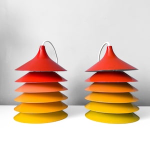 Op de afbeelding: Twee retro lampen met een gelaagd, conisch ontwerp. Elke lamp heeft een kleurengradient, van rood aan de bovenkant naar geel aan de onderkant. De lampen zijn verbonden met een wit snoer, wat suggereert dat het hanglampen zijn.