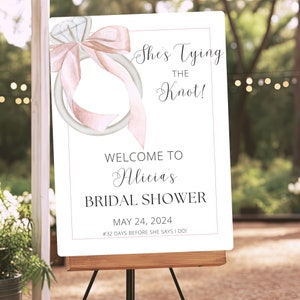 May include: A white sign with a pink and white watercolor illustration of a ring with a bow. The text on the sign reads "She's Tying the Knot!"  "Welcome to Alicia's Bridal Shower"  "May 24, 2024"  "#32 Days Before She Says I Do!"