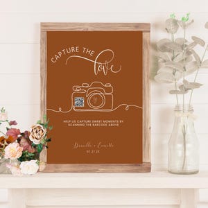 Puede incluir: Un letrero enmarcado con fondo marrón y texto blanco. El texto dice "Capture the love" encima de un dibujo lineal de una cámara. Debajo de la cámara está el texto "Ayúdanos a capturar momentos dulces escaneando el código de barras de arriba."