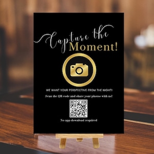 May include: A black sign with gold text and a gold camera icon. The sign reads "Capture the Moment!" and includes a QR code and the text "We want your perspective from the night! Scan the QR code and share your photos with us! No app download required."