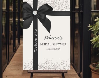 May include: A welcome sign for a bridal shower. The sign features a black bow and ribbon, with the words "Welcome to" and "Rebecca's Bridal Shower" in elegant script. The background is white with scattered pearl accents.