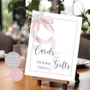 May include: A white sign with a pink watercolor illustration of a ring with a diamond and a bow. The sign says "Cards and Gifts Thank You!" with a pink heart.