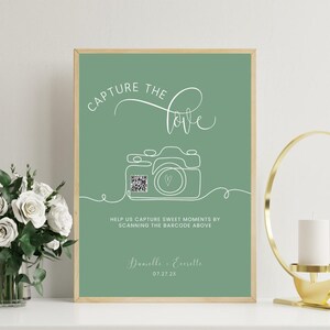Könnte beinhalten: Ein gerahmtes, salbeigrünes Schild mit den Worten "CAPTURE the love" in weißer Schrift. Eine Strichzeichnung einer Kamera mit einem QR-Code befindet sich unter dem Text. Auf dem Schild steht "HELFT UNS, SCHÖNE MOMENTE ZU ERFASSEN, INDEM IHR DEN BARCODE OBEN SCANNT".