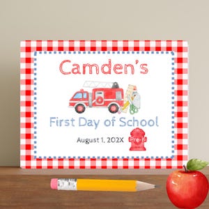 May include: A rectangular sign with a red and white checkered border. The sign features the text "Camden's First Day of School" with a red fire truck illustration. A pencil and a red apple are on the wooden surface.