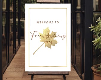 May include: A white Friendsgiving 2024 sign with gold lettering and a gold leaf illustration, displayed on a wooden easel. The sign reads "Welcome to Friendsgiving". The sign is framed with a thin gold border.