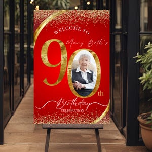 May include: A red and gold 90th birthday celebration sign. The sign features the text "Welcome to Mary Beth's" and "Birthday Celebration". A photo of a smiling elderly woman is in the center. The sign is on a wooden easel.