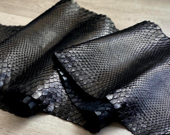 May include: A long, flexible strip of black snakeskin leather featuring a distinct, repeating scale pattern across its entire surface. The material is folded in several places, showcasing the texture and dark, reflective finish of the scales.