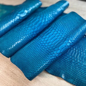 May include: A vibrant teal blue snakeskin leather hide featuring a glossy finish and a distinct, repeating scale pattern. The material is folded in multiple layers, showcasing the texture and the deep, saturated color across the entire surface.