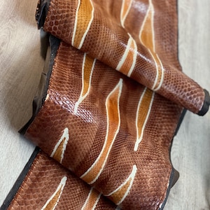 May include: A long, rectangular strip of textured snakeskin leather featuring a repeating pattern of brown scales accented by elongated, diagonal stripes in shades of golden yellow and white. The material has a glossy finish and dark edges.
