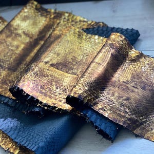 Op de afbeelding: Close-up van verschillende stukken goud- en bruin slangenleer. Het leer heeft een metallic glans en een textuur, geschubd patroon. De randen van het leer zijn donker en de stukken zijn op elkaar gelegd.