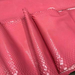 May include: A sheet of bright pink snakeskin leather featuring a distinct, raised scale pattern across the entire surface. The material is folded over itself, showcasing the texture and the vibrant, uniform pink coloration of the reptile hide.