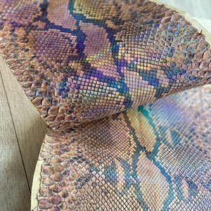 May include: A piece of snakeskin leather featuring a textured scale pattern with an iridescent finish. The material displays a gradient of colors including metallic copper, bronze, deep purple, and teal blue, creating a shifting, multi-tonal effect.