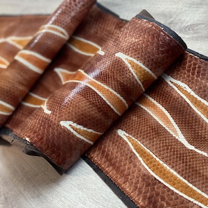 May include: A long, rectangular strip of textured brown snakeskin leather featuring a repeating pattern of diagonal, cream-colored stripes. The material has a distinct, fine-scaled surface and is finished with a dark, solid-colored edge trim.
