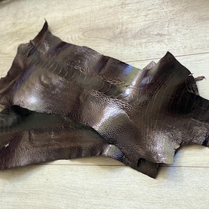 May include: Two pieces of dark brown ostrich leather with a textured surface. The leather has a glossy finish and is arranged on a light wood grain surface.