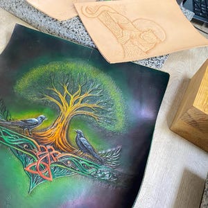 May include: Hand-tooled leather art piece featuring a tree with two crows, Celtic knot, and vibrant colours. The artwork is on a dark green background with a gradient effect. The tree has a yellow trunk and green foliage.