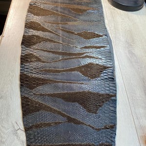 Peut inclure: Une longue bande étroite de cuir de peau de serpent brun foncé et noir présentant un motif d'écailles distinct et répétitif. La peau est posée à plat, révélant une surface texturée avec des motifs en losange alternant brun foncé et clair.
