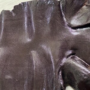 Pode incluir: Uma peça de couro castanho escuro texturizado com um padrão de escamas fino e uniforme que imita a aparência da pele de lagarto. O material tem um acabamento brilhante com reflexos subtis de luz sobre a sua superfície complexa.