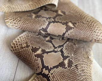 May include: A piece of natural snakeskin leather featuring a repeating diamond-shaped pattern in shades of tan, beige, and dark brown. The material has a textured, scaly surface with a visible grain and a flexible, folded appearance throughout the piece.