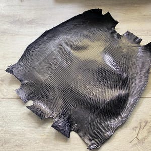 Może przedstawiać: Kawałek czarnej skóry o fakturze jaszczurki z metalicznym połyskiem. Skóra ma wzór łusek i nieregularne krawędzie, co sugeruje, że jest to skóra. Materiał ten jest prawdopodobnie używany do tworzenia przedmiotów takich jak portfele lub torby.