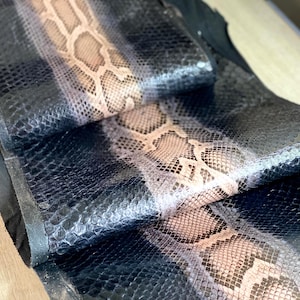 Op de afbeelding: Een strook gestructureerd slangenleer met een centrale band van beige en bruine schubben, geflankeerd door diepzwarte schubben aan beide zijden. Het materiaal heeft een duidelijk reptielpatroon met een glanzende afwerking en is dubbelgevouwen.