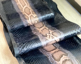 May include: A strip of textured snakeskin leather featuring a central band of tan and brown scales flanked by deep black scales on both sides. The material has a distinct reptilian pattern with a glossy finish and is folded over itself.