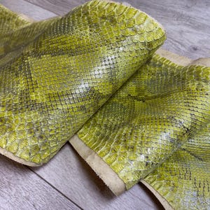 May include: A long, flexible strip of genuine snakeskin leather featuring a vibrant chartreuse and lime green colour palette. The material displays a distinct, repeating diamond-shaped scale pattern with dark grey accents between the individual scales.