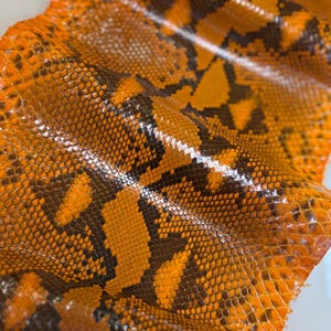 May include: A sheet of orange and dark brown snakeskin leather featuring a repeating, textured scale pattern. The material has a glossy finish and is folded in soft, undulating waves, highlighting the intricate detail of the reptile skin.