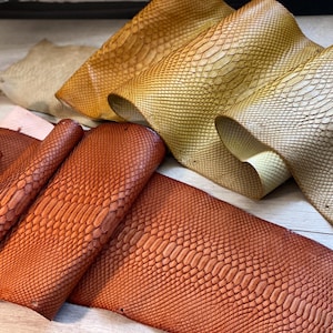 May include: An assortment of textured leather hides in various colors, including shades of orange, tan, and cream. The leather has a scale-like pattern, suggesting a reptile skin design. The hides are arranged in overlapping layers, showcasing their different textures and hues.