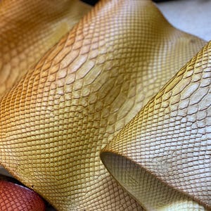 May include: A collection of folded leather sheets featuring a distinct, embossed snakeskin pattern. The material displays a gradient of warm golden-yellow and tan tones, with a textured surface that mimics the appearance of overlapping reptile scales.