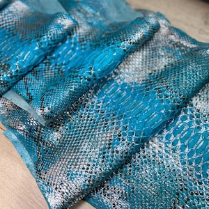 May include: A piece of genuine snakeskin leather featuring a vibrant turquoise and metallic silver finish. The material displays a distinct, repeating scale pattern with dark grey accents throughout the textured surface of the hide.