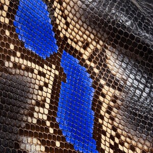 May include: A textured material featuring a repeating snakeskin pattern with scales in shades of deep black, natural beige, and vibrant royal blue. The surface displays a detailed, organic scale structure with a distinct color gradient.