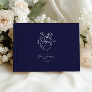May include: A navy blue wedding guest book with a white heart-shaped locket design and the names "the Jasons" in script. The book also includes the text "WEDDING GUEST BOOK" and the wedding date.