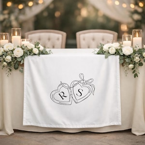 May include: White table runner with a black line drawing of two connected hearts, each with a letter inside. The heart on the left has an "R" and the heart on the right has an "S". The runner is on a table with floral arrangements and candles.