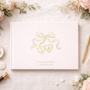 May include: A light pink wedding guest book with gold foil accents, featuring a bow and heart design. The book is surrounded by flowers, a pen, and wedding rings. The text on the book reads "WEDDING GUEST BOOK JANUARY 25, 2026."
