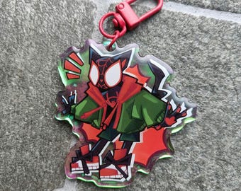 May include: A clear acrylic keychain featuring a cartoon character in a green jacket and red accents. The keychain has a red clasp and a unique, jagged edge design. The character has white eyes and a black mask. The overall design is vibrant and eye-catching.