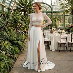 May include: A two-piece white dress featuring a long-sleeved lace crop top with a sweetheart neckline and a floor-length skirt with a high side slit. The garment is adorned with intricate floral lace detailing on the bodice and sleeves.