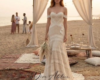 May include: A floor-length off-the-shoulder wedding dress featuring intricate cream-colored lace patterns, a fitted bodice, and a flared skirt. The garment includes short lace sleeves and a delicate waist tie with tassels, creating a textured look.