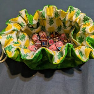 May include: A drawstring dice bag filled with various polyhedral dice. The bag has a bright green ruffled edge and a white interior patterned with yellow pineapples. The dice are in various colours, including pink, red, and blue.