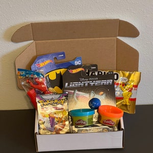 May include: An open cardboard box filled with various toys and treats. The box contains a red and blue Batman toy car, a red plastic water gun, a pack of Pokemon trading cards, two small containers of Play-Doh, a Buzz Lightyear toy, and a bag of gummy bears.