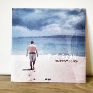 Puede incluir: Una portada de disco de vinilo con un fondo de acuarela azul y blanco. La portada presenta a un hombre caminando por una playa con el texto "Lyric Reborn Balcony Beach" impreso en la parte inferior.