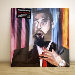 May include: Album cover art featuring a painting of a man with a beard and a suit. The background is a colorful abstract design. The album title is "Your Old Droog" and the artist is "Rodney Dangerfield".