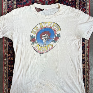 May include: White t-shirt with a distressed, vintage look. The shirt features a circular graphic of a skull with a red bandana and roses, with the text "Grateful Dead" in a psychedelic font.