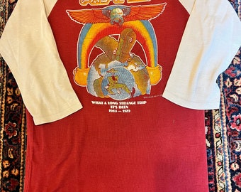 May include: A red and cream-colored baseball style shirt with the words "Grateful Dead" and a graphic of a rainbow, earth, and figures. The text "What a long strange trip it's been 1965-1979" is also printed on the shirt.