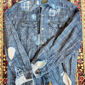 Puede incluir: Una camisa vaquera azul con estampado de cuadros y parches. La camisa tiene un cierre de botones en la parte delantera y mangas largas. La etiqueta de la camisa dice "KOUNTRY".