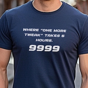 May include: A navy blue t-shirt with white text that reads "WHERE 'ONE MORE TWEAK' TAKES 6 HOURS. 9999." The shirt is short-sleeved and worn by a person smiling in an outdoor setting.